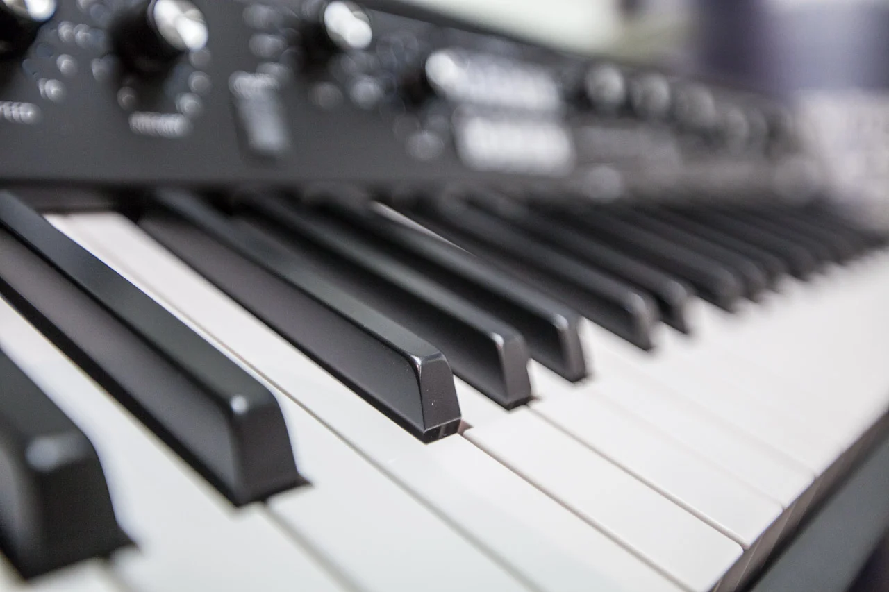 Close-up of a keyboard synthesizer's black and white keys with control knobs blurred in the background; monochrome tones.