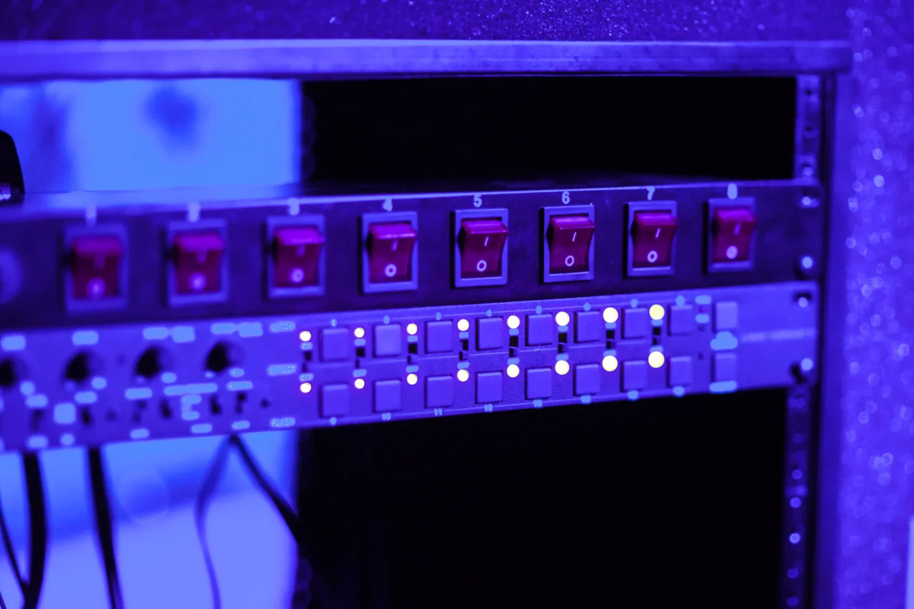 Close-up of an equipment rack with red power switches and small glowing indicator lights, bathed in purple light; technical music hardware